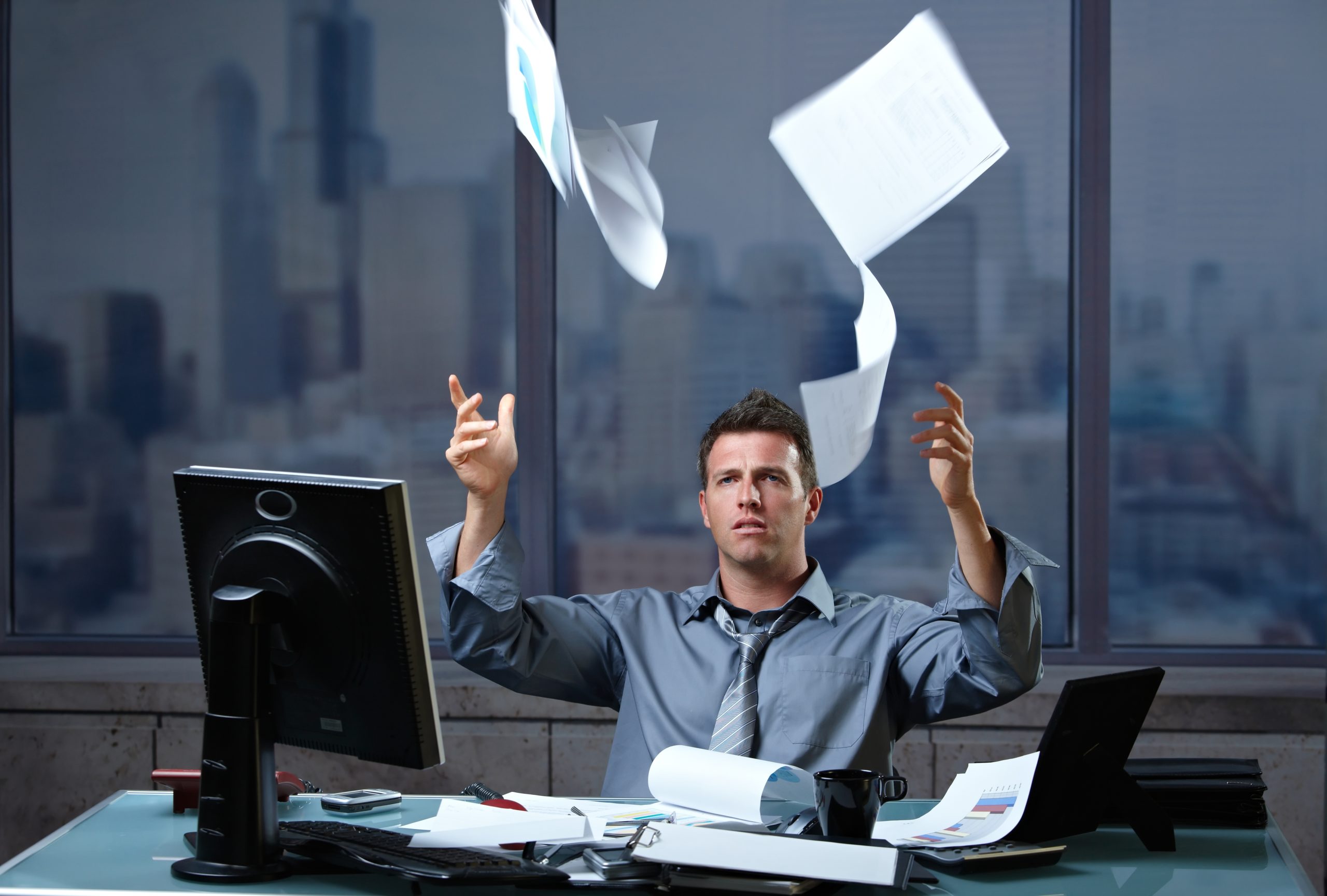 Exhausted professinal throwing documents into air sitting at office desk in overtime.
